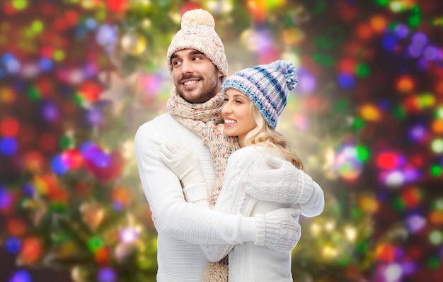 inverno, moda, casal, natal e conceito de pessoas - sorrindo homem e mulher em chapéus e cachecol abraçando sobre fundo de luzes