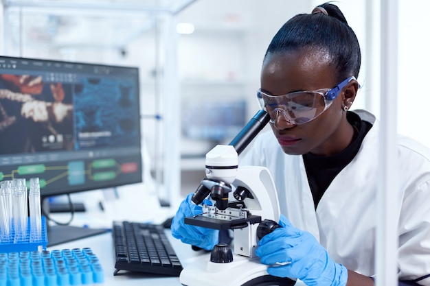 Investigador médico de etnia africana con microscopio que investiga y evalúa la muestra. Científico de la salud negro en el laboratorio de bioquímica con equipo estéril.