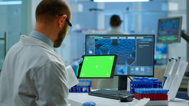 Investigador sosteniendo y mirando la tableta con pantalla chroma key en un moderno laboratorio equipado. Equipo de microbiólogos que realizan investigación de vacunas escribiendo en un dispositivo con pantalla verde, pantalla de maqueta aislada.