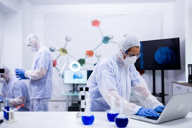 Foto investigadora estilizando en una computadora portátil con una solución azul humeante de tubos de ensayo frente a ella. científico en laboratorio moderno.