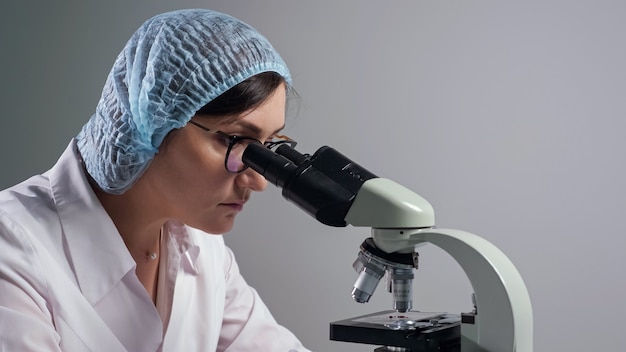 Investigadora con gafas en bata blanca y gorra azul mira la muestra líquida a través del microscopio contemporáneo en el lugar de trabajo en el laboratorio de la clínica closeup