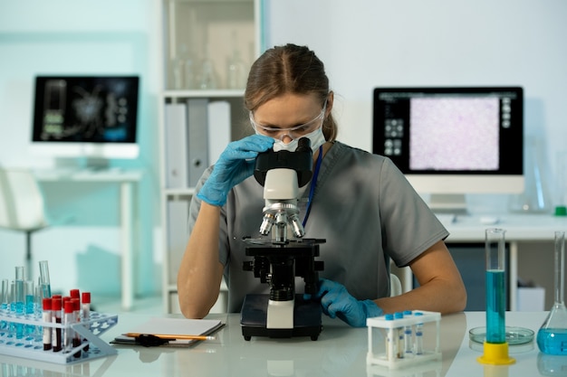 Investigadora joven ocupada en guantes y gafas sentado en la mesa con soporte para tubos de ensayo y trabajando con muestras en el laboratorio