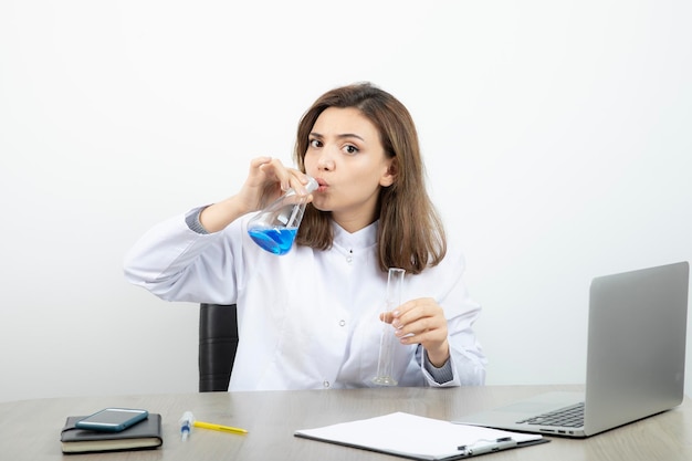 Investigadora de laboratorio sentada en el escritorio y sosteniendo una botella de vidrio médico con líquido azul. foto de alta calidad