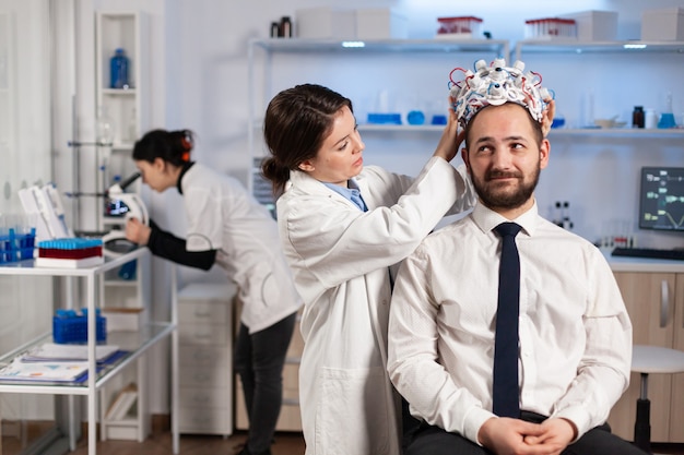 Investigadora poniendo auriculares de exploración de ondas cerebrales al paciente que desarrolla experimentos utilizando monitores de laboratorio de estudio cerebral. Hombre sentado en una silla mientras el científico dispositivo de ajuste examinando el sistema nervioso