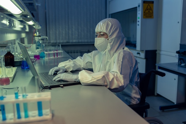 Foto investigadora en ropa de trabajo protectora sentada en la mesa y escribiendo en la computadora portátil durante su trabajo en el laboratorio