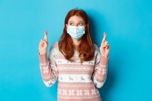 Invierno, coronavirus y concepto de distanciamiento social. Linda chica esperanzada con el pelo rojo, con mascarilla, cruzar los dedos y pedir deseos, de pie sobre fondo azul.