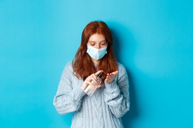 Invierno, covid-19 y concepto de distanciamiento social. Estudiante pelirroja en mascarilla limpia las manos con desinfectante, con antiséptico, de pie contra el fondo azul.