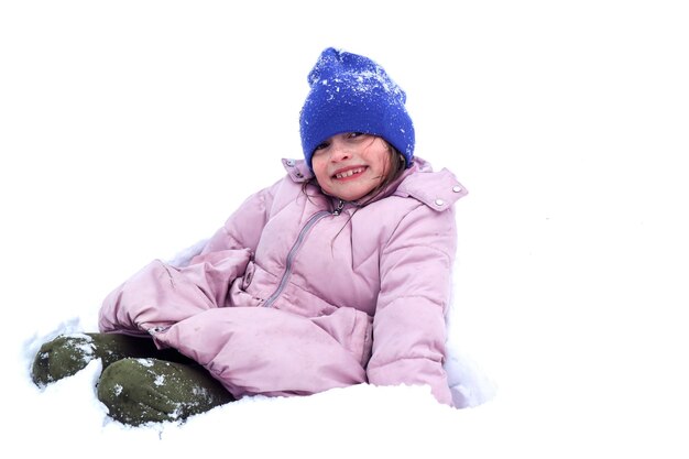 El invierno es una época de juegos y placeres, una niña sonriente de ocho años con una chaqueta pastel y un sombrero azul se sienta en el primer plano de la nieve