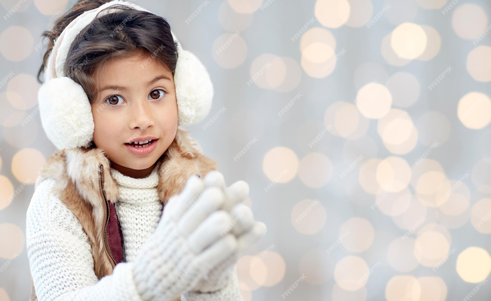 Invierno, gente, concepto de felicidad navideña - niña feliz con orejeras y  guantes durante las vacaciones luces de fondo