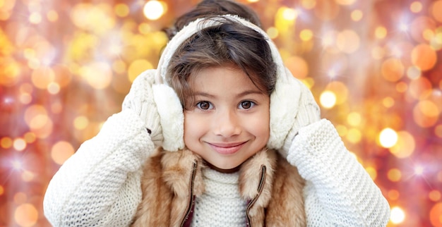 Invierno, gente, navidad y concepto de ropa - niña feliz usando orejeras y  guantes sobre fondo de luces de vacaciones doradas