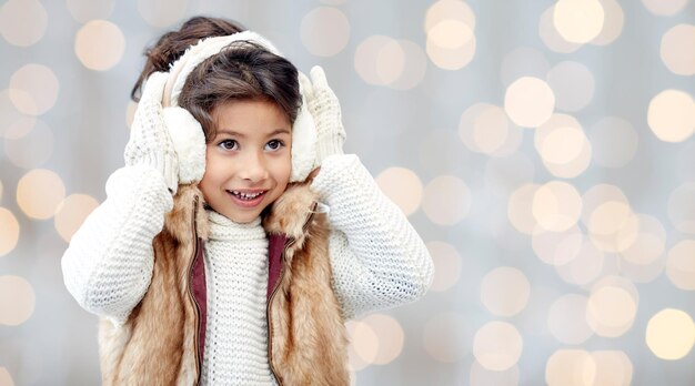 Foto invierno, gente, navidad, moda y concepto de infancia - niña feliz usando orejeras y guantes durante las vacaciones luces de fondo