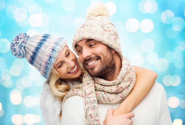 invierno, moda, pareja, navidad y concepto de la gente - hombre y mujer sonrientes con sombreros y bufanda abrazándose sobre el fondo azul de las luces navideñas