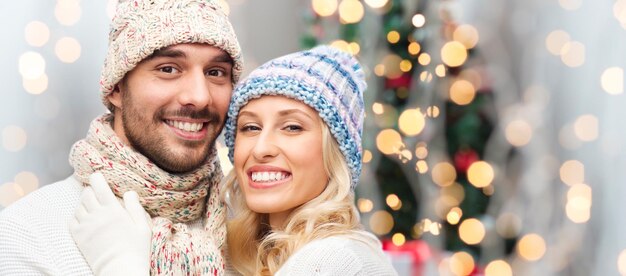 invierno, moda, pareja, navidad y concepto de la gente - hombre y mujer sonrientes con sombreros y bufanda abrazándose sobre el fondo de las luces navideñas