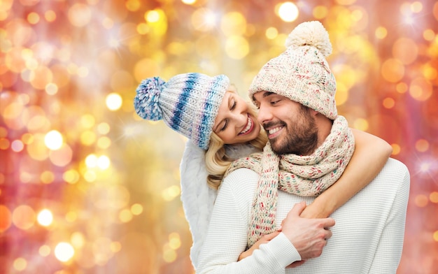 invierno, moda, pareja, navidad y concepto de la gente - hombre y mujer sonrientes con sombreros y bufanda abrazándose sobre el fondo de las luces