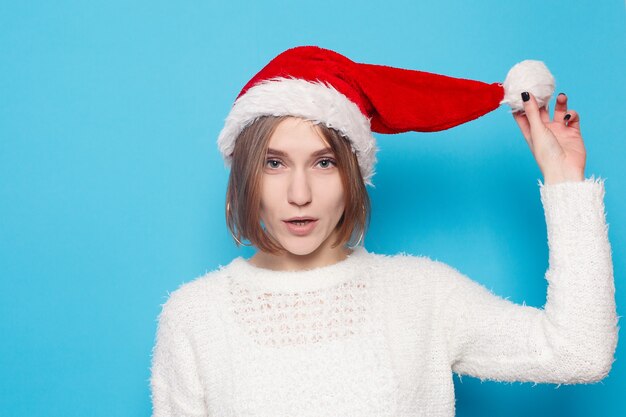 Invierno, navidad, gente, concepto de belleza - bonita rubia con sombrero de navidad sobre superficie azul claro