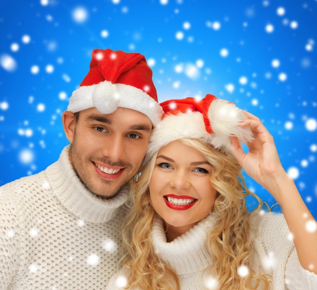 invierno, vacaciones, navidad y concepto de la gente - pareja sonriente en suéteres y sombreros de ayudante de santa sobre fondo azul nevado