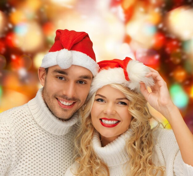 invierno, vacaciones, navidad y concepto de la gente - pareja sonriente en suéteres y sombreros de ayudante de santa sobre fondo de luces rojas