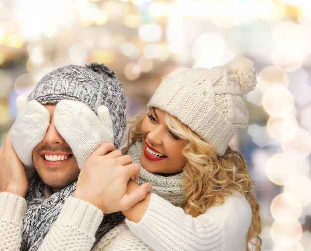 invierno, vacaciones, navidad y concepto de la gente - pareja sonriente en suéteres y sombreros de ayudante de santa sobre fondo de luces