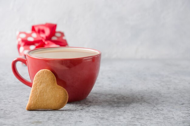 Invitación de San Valentín. Rojo taza de café con leche, regalo y galletas de corazón en gris. De cerca.