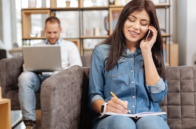 Involucrado en el trabajo. Encantado de mujer bonita agradable hablando por teléfono y tomando notas mientras está sentado en el sillón