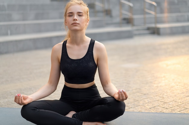 Ioga e meditação na cidade moderna Mulher caucasiana relaxa posição de lótus Sentar-se Tapete de ioga ao ar livre Parque de verão em piso de concreto Jovem adulto apto feminino relaxando após o treino de ioga Siddhasana