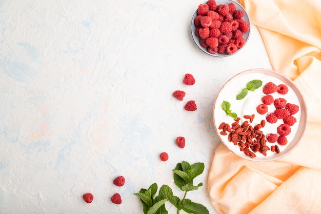 Iogurte com framboesa e goji berries em uma tigela de cerâmica na superfície de concreto branco e tecido de linho laranja