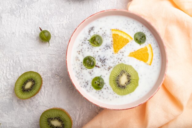 Iogurte com kiwi, groselha, chia em uma tigela de cerâmica sobre fundo cinza de concreto e têxteis de linho laranja. vista de cima, postura plana, close-up.