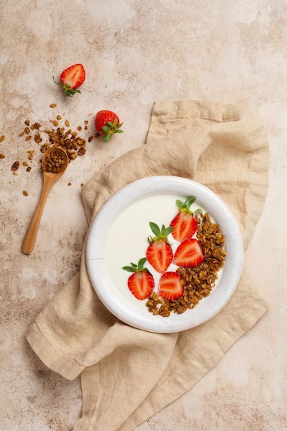 Iogurte grego em uma tigela branca com ingredientes para fazer granola de café da manhã e morangos frescos na velha mesa bege. Vista do topo