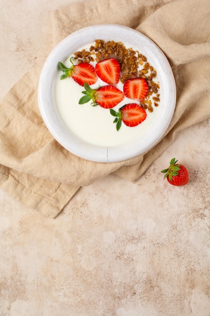 Iogurte grego em uma tigela branca com ingredientes para fazer granola de café da manhã e morangos frescos na velha mesa bege. Vista do topo.