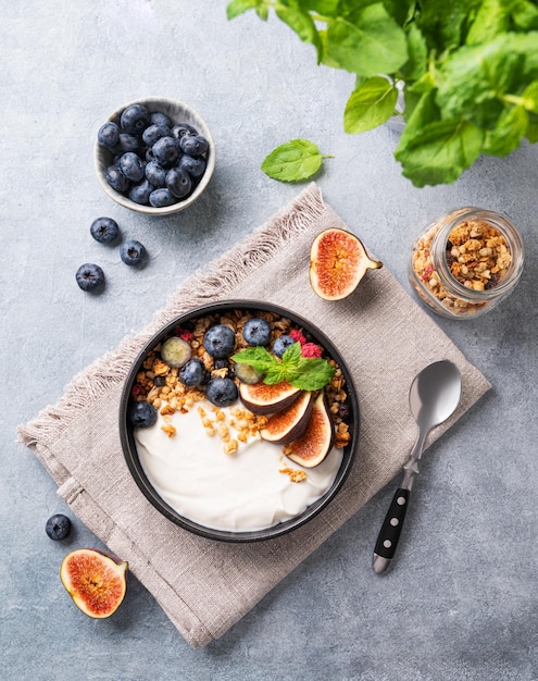 Iogurte natural com bagas de muesli e figos numa tigela preta sobre fundo azul com menta Pequeno-almoço saudável e nutritivo