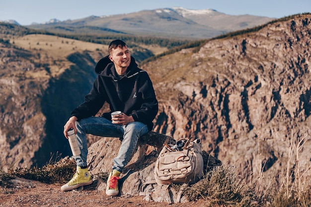 Foto ir a tomar una bebida caliente. un chico joven está sentado en una roca con una taza de té y un termo para viajar en sus manos. mirada desviada hacia un lado y una sonrisa pacífica. fondo de montañas.