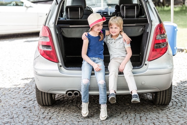 Irmã mais nova e irmão sentado no porta-malas de um carro com malas. o conceito de viagens em família