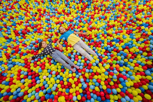 Irmão com irmã brincando no parquinho do centro de jogos interno Menina com menino deitado em bolas coloridas na piscina de bolas