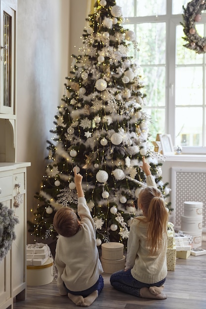 Irmão e irmã mais novos se abraçam e olham para uma linda árvore de natal decorada com deleite e sonhos, fazem desejos