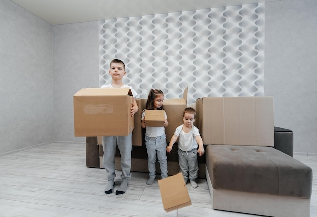 Irmãos e irmã estão brincando e gostando de se mudar para um novo apartamento no contexto de caixas de papelão e um sofá compra de imóveis entrega de inauguração