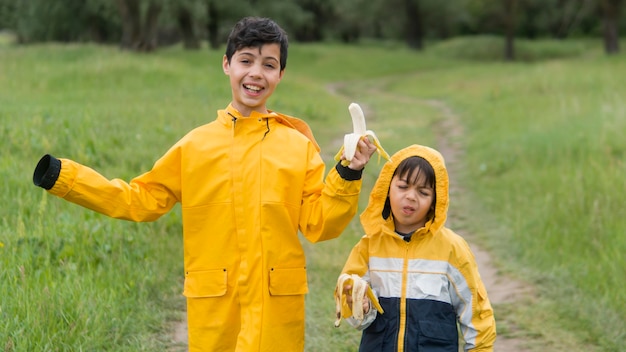 Foto irmãos em capa de chuva comendo bananas plano médio