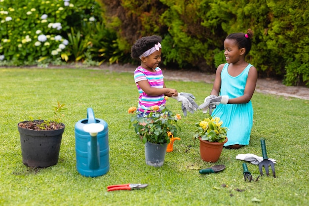 Irmãs afro-americanas sorridentes e brincalhonas ajoelhadas enquanto usavam luvas perto de plantas no jardim