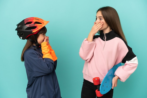 Irmãs caçulas praticando ciclismo e skatista isoladas em um fundo azul cobrindo a boca com as mãos por dizer algo impróprio