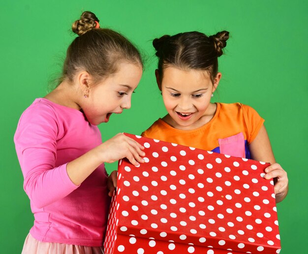 Irmãs com caixa de presente pontilhada de bolinhas vermelhas para férias. Meninas com rostos sorridentes curiosos posam com presentes sobre fundo verde. As crianças abrem o presente para o Natal. Ano novo apresenta o conceito.