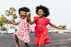 Irmãs gêmeas afro correndo na praia enquanto brincava com o avião de brinquedo de madeira - estilo de vida da juventude e conceito de viagens - foco principal no rosto direito do garoto