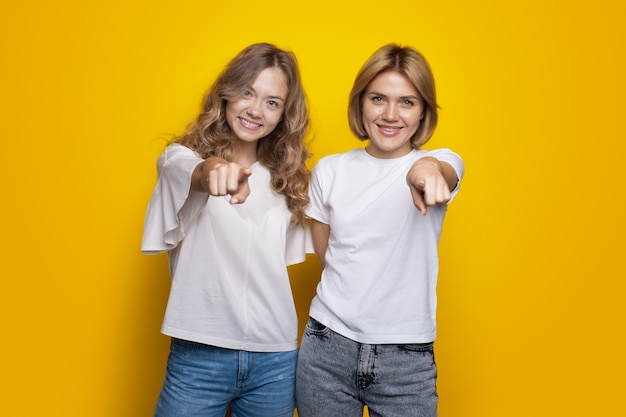 Irmãs loiras apontando para a câmera sorrindo na parede amarela do estúdio vestindo camiseta e jeans