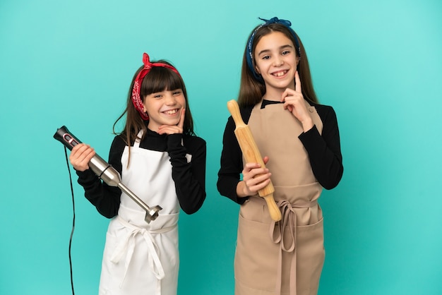 Irmãs mais novas cozinhando em casa isoladas em um fundo azul, sorrindo com uma expressão doce