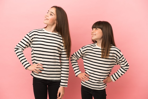 Irmãs mais novas isoladas em um fundo rosa posando com os braços na cintura e rindo