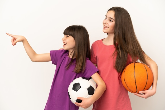 Irmãs mais novas jogando futebol e basquete isoladas em um fundo branco apontando para o lado para apresentar um produto