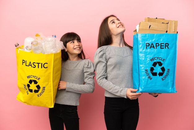 Irmãs mais novas reciclando papel e plástico isolados em um fundo rosa olhando para cima enquanto sorriem