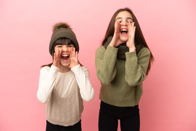 Irmãs mais novas vestindo roupas de inverno isoladas em um fundo rosa gritando e anunciando algo