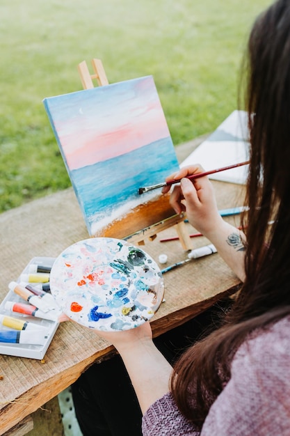 Irreconhecível jovem artista feminina pintando seu próprio cenário de praia com óleo sobre uma tela ao ar livre. Amat