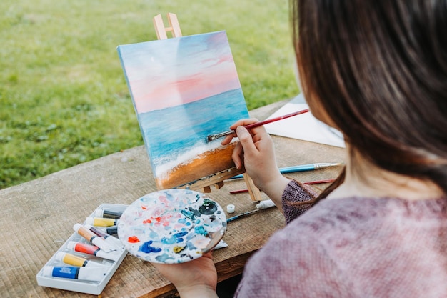 Irreconhecível jovem artista feminina pintando seu próprio cenário de praia com óleo sobre uma tela ao ar livre.