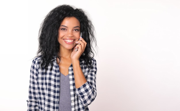 Irresistible chica afroamericana en camisa de franela sostiene un teléfono inteligente escucha su oído izquierdo, sonriendo y mirando directamente a la cámara