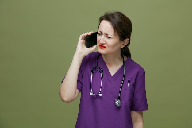 Irritada doctora de mediana edad con uniforme y estetoscopio alrededor del cuello hablando por teléfono mirando al lado aislado en el fondo verde oliva